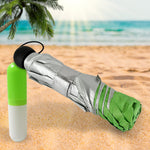 A compact green and silver umbrella is folded next to its capsule case on a sandy beach, with a palm tree and ocean in the