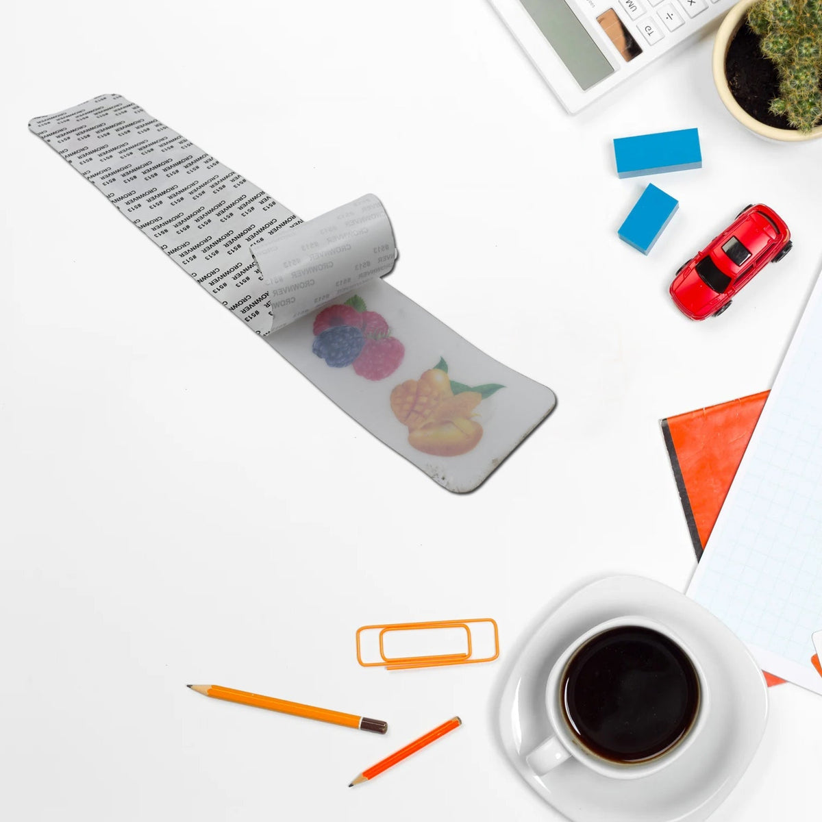 A white adhesive wall hook strip with fruit designs is shown peeling off its backing on a white desk with office supplies.