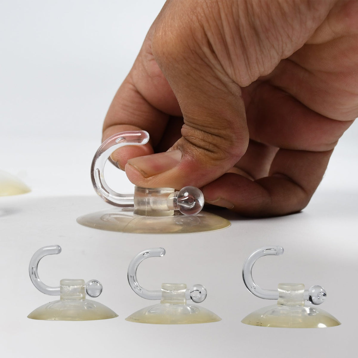 A hand demonstrates how to attach a transparent suction cup hook, with three more hooks on a white surface.