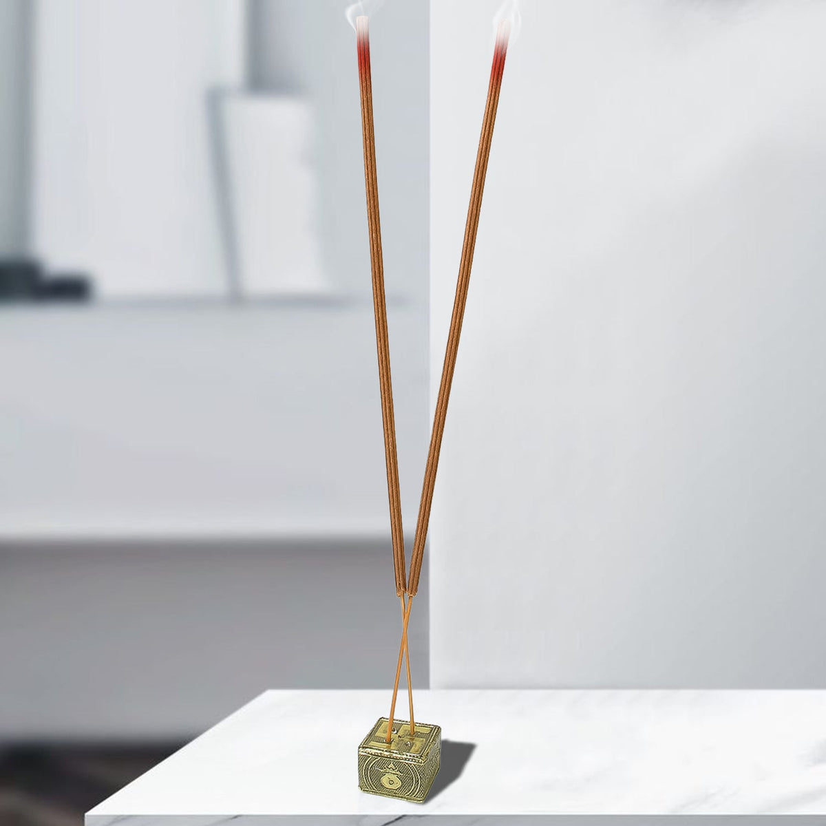 Two burning brown incense sticks in a golden holder on a white marble surface in a bright room.