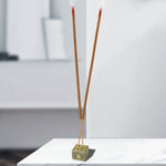 Two burning brown incense sticks in a golden holder on a white marble surface in a bright room.