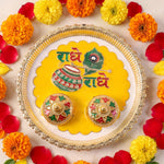 A silver and yellow Radhe Radhe pooja thali set with two small decorative containers, surrounded by marigolds and rose