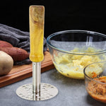 A stainless steel masher with a light wooden handle stands beside bowls of mashed potatoes and sweet potatoes.