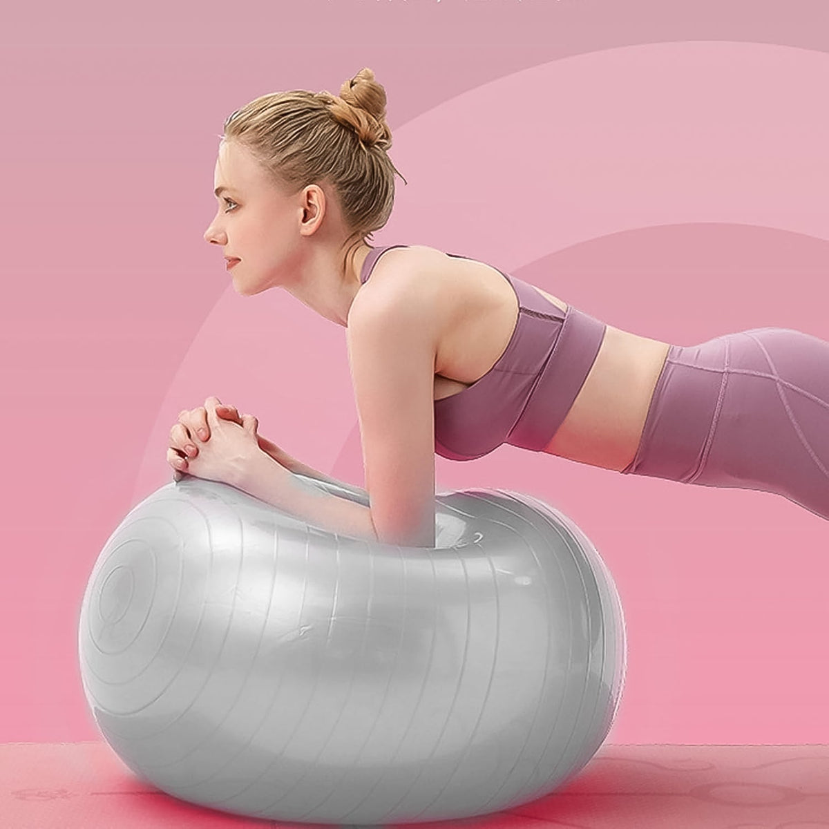 A woman in a purple sports bra and leggings holds a plank position with her forearms on a grey stability ball, against a