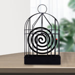 A black iron birdcage-style holder with a burning mosquito coil inside sits on a light wooden table.