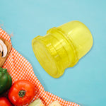 A yellow plastic manual food grinder is angled on a blue background, with a red and white checkered cloth and vegetables in