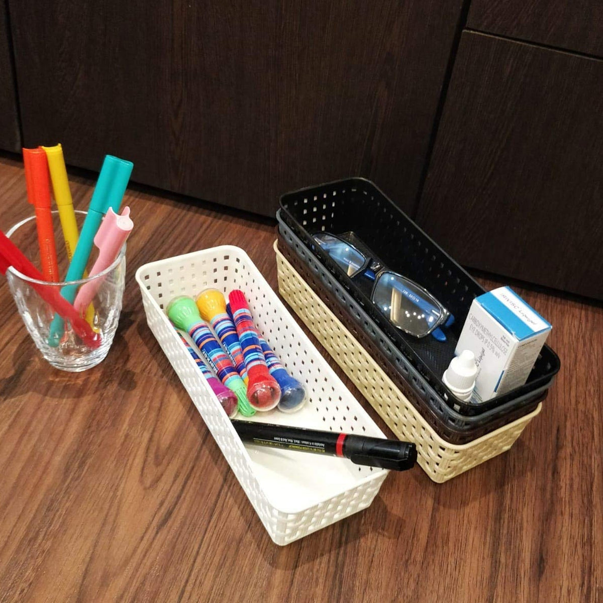 Two plastic baskets and a glass of markers on a wooden floor next to a dark cabinet.