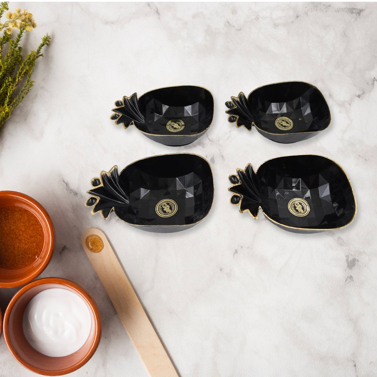 Four black ceramic pineapple-shaped snack bowls with gold accents on a white marble surface next to small bowls of