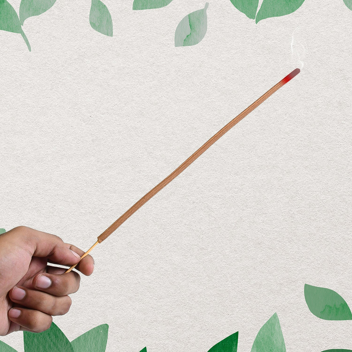 A hand holding a single burning brown incense stick against a light background with green leaf accents.