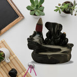 A black Ganesha backflow incense burner with smoke, succulents, and a small mat on a white surface.