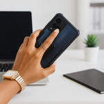 A hand holding a black smartphone with a silver ring stand, with a laptop and notebook in the background.