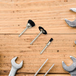 Three black nylon brushes with silver shafts are arranged on a light wooden surface surrounded by silver wrenches.