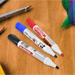 Three whiteboard markers in black, blue, and red, with their caps on, lying on a wooden surface with drafting tools in the