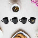 Four black ceramic tea cups with gold accents on a white marble surface next to a glass infuser filled with loose leaf tea.