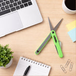 Green pen-shaped scissors and a black safety cutter lie on a wooden desk next to a laptop, notebook, and coffee.