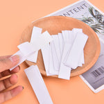 A hand holds a white strip of tape over a wooden bowl filled with many similar strips, next to a magazine.