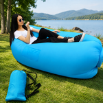 A woman in sunglasses relaxes on a blue inflatable lounger by a lake, with a matching carry bag on the grass.