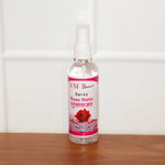 A clear bottle of SM Beauty rose water spray with a pink label stands on a wooden surface against a white tiled wall.