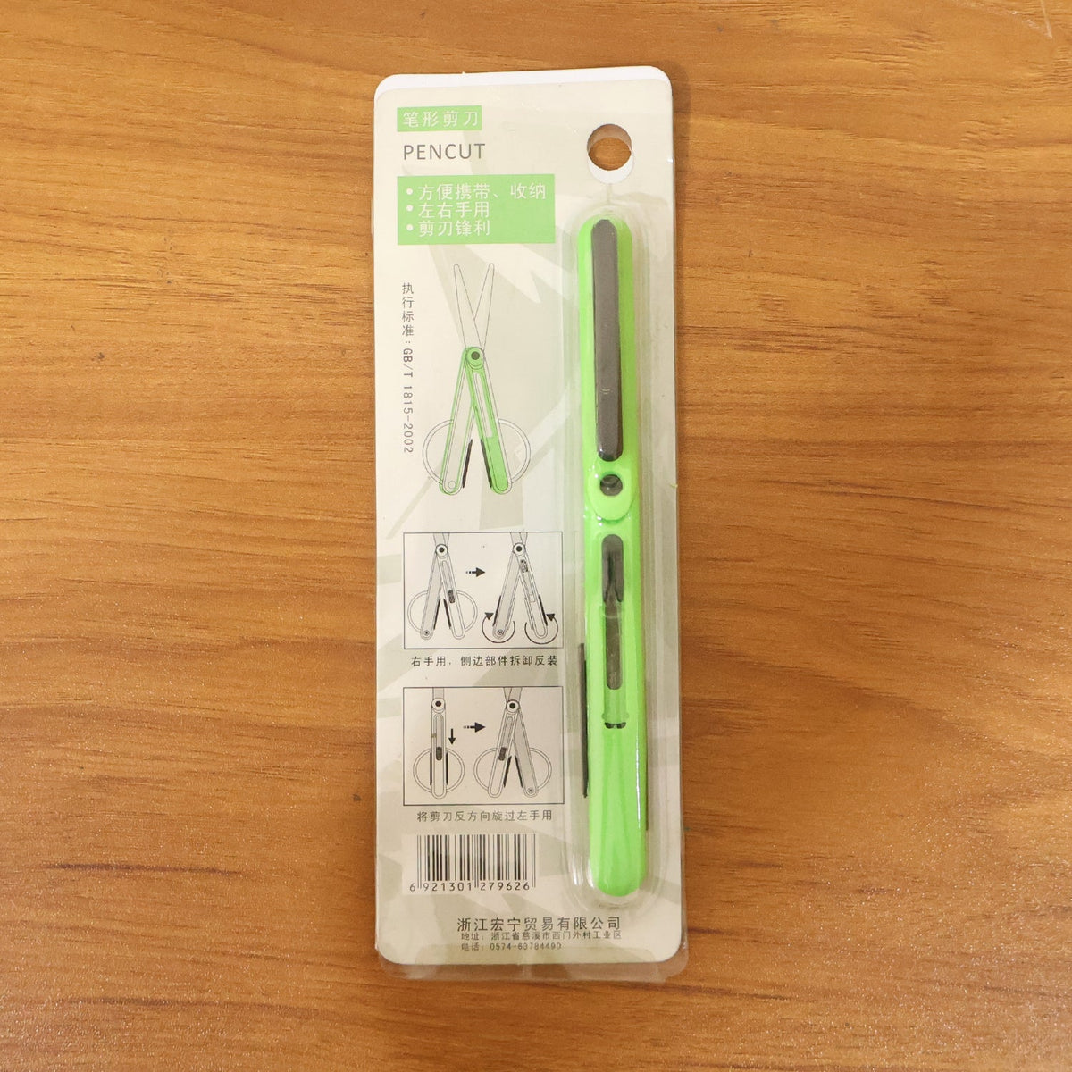 The green pen-shaped safety cutter is displayed in its retail packaging on a wooden surface.