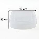 A clear plastic sandwich container with its lid is on a striped cloth on a wooden table with fruit and vegetables.