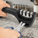 Hands hold a black three-stage sharpener while sharpening blue-handled scissors on a light speckled countertop.