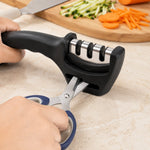 Hands hold a black three-stage sharpener, sharpening blue-handled scissors with chopped vegetables in the background.