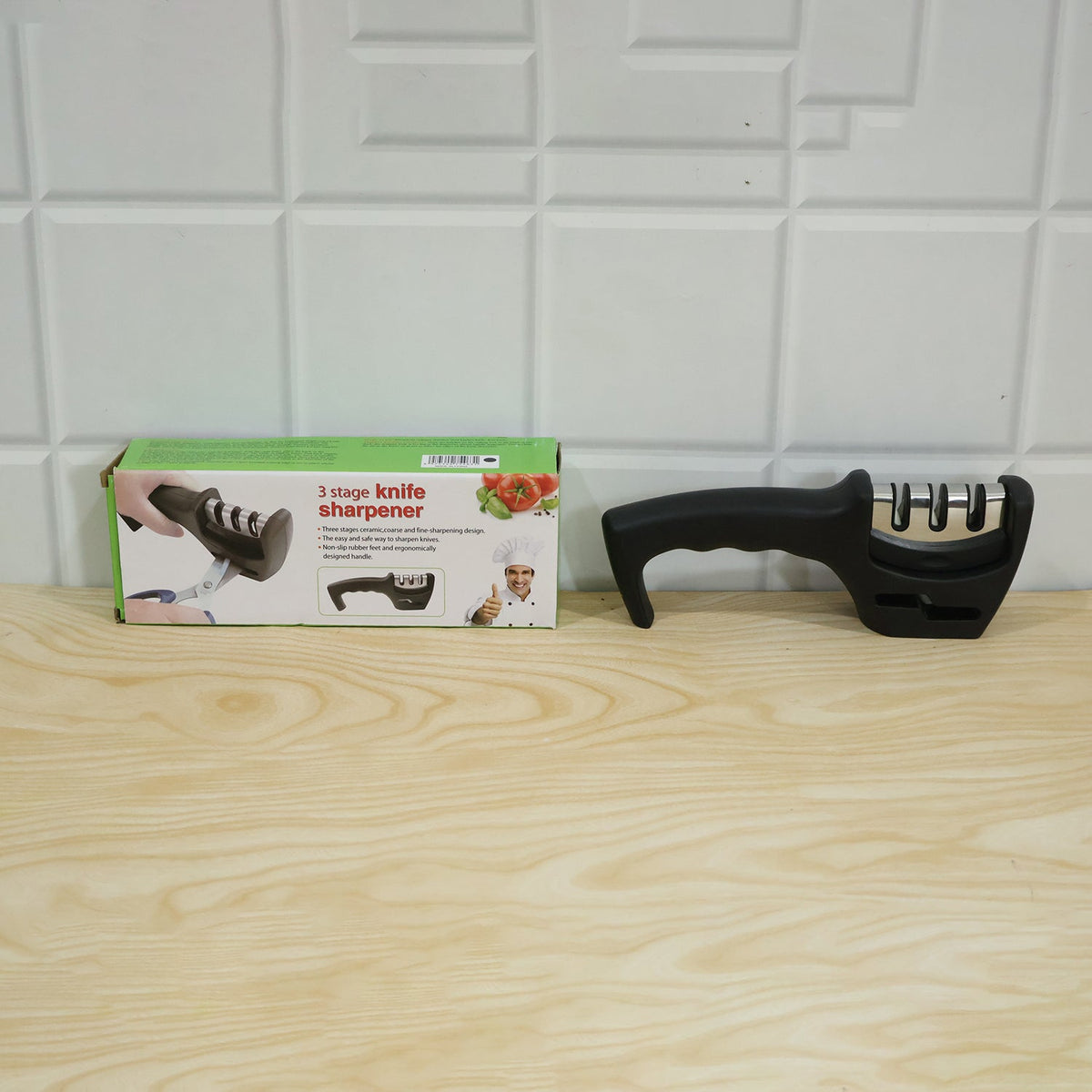 A black three-stage knife sharpener and its product box are placed side-by-side on a light wooden surface.