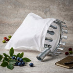 The fruit picker's white bag and metal prongs are on a textured grey surface, surrounded by blueberries, leaves, and a
