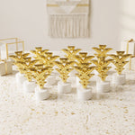 A dozen golden Christmas tree LED tealights stand on a white speckled surface, surrounded by gift boxes.