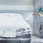 A person's hands hold a teal pressure washer, spraying water onto a white foamy car in front of a gray garage door.
