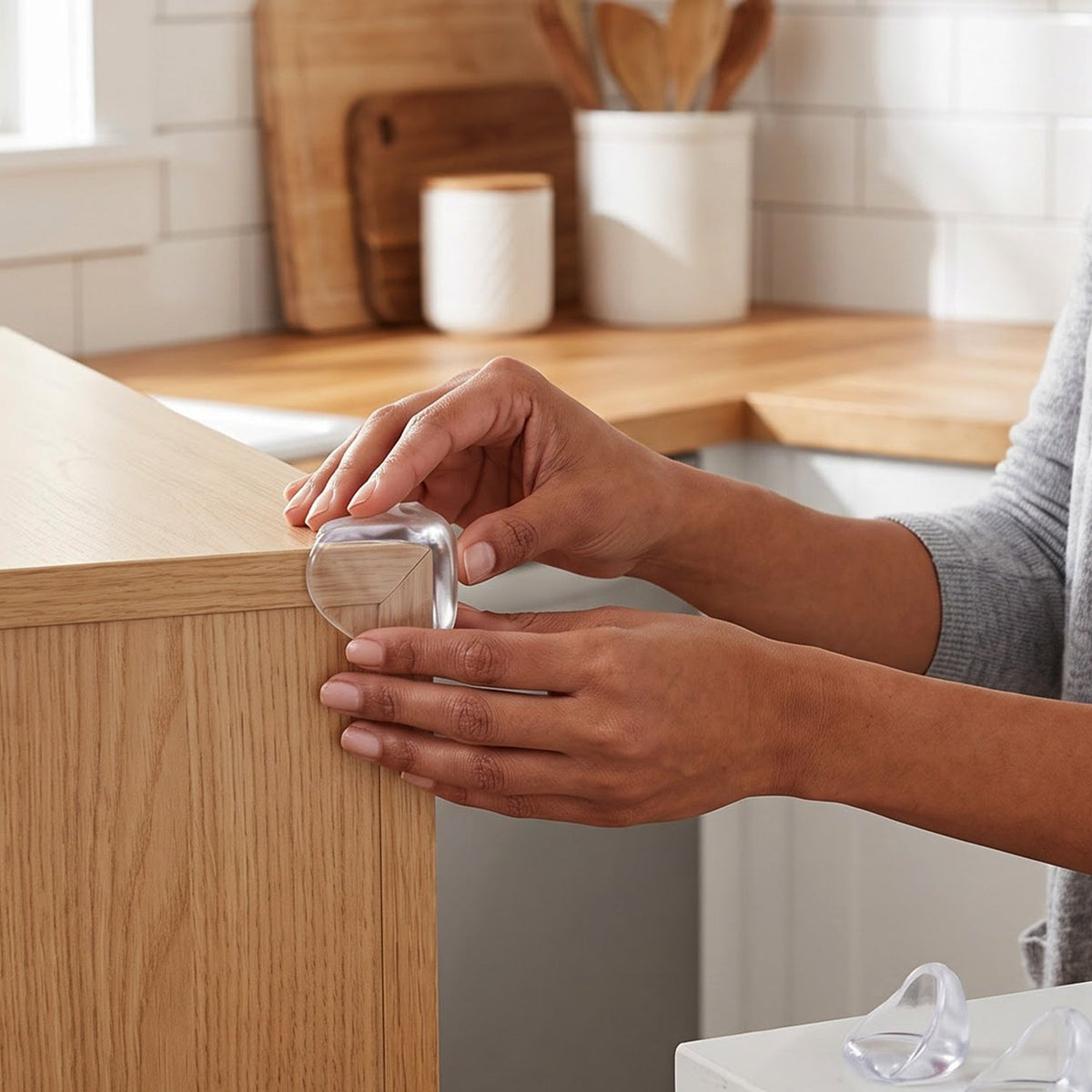 A person's hands applying a clear, rounded corner protector to a light wood table edge in a kitchen.