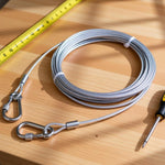 A coiled stainless steel wire rope with two locking hooks on a wooden surface next to a yellow measuring tape and a