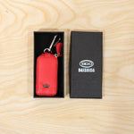 Red leather car key case with a silver snap hook, placed inside a black gift box next to a branded black box.