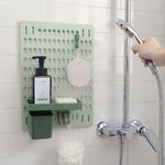 A light green pegboard with a dark green shelf holding soap and lotion, a dark green cup, and hooks holding a loofah, in a