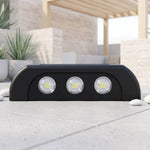 A black solar-powered LED wall light with three bright white LEDs on a light grey tiled patio with white pebbles and plants.