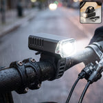 Black rectangular bicycle headlight with a clear lens, mounted on handlebars, shining brightly in the rain at night.