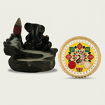 A black Ganesha backflow incense burner with smoke and a yellow and gold puja thali with peacock design on a white