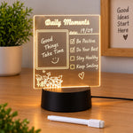 A glowing acrylic message board lamp on a wooden desk, displaying a daily moments list with a positive message.