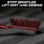 A close-up of the red and white handled brush with brown bristles on a dark textured surface, with text about stiff bristles.