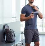 A man in a blue t-shirt and grey shorts sprays a blue perfume bottle onto his neck, standing next to a suitcase and backpack.