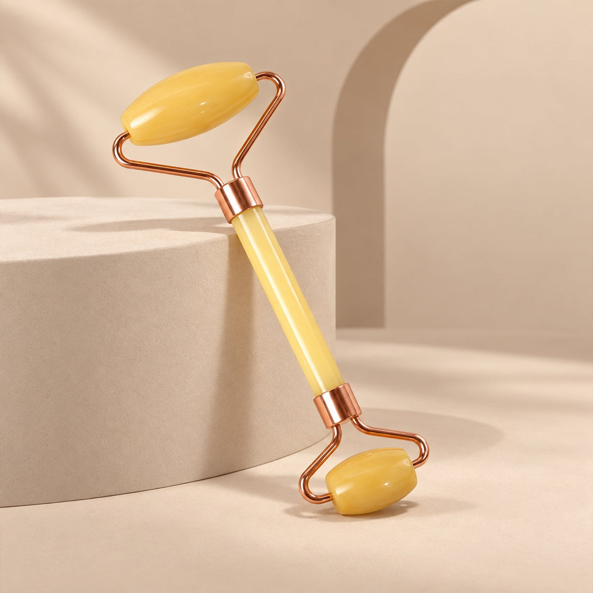 A yellow jade facial roller with rose-gold frame is angled on a light beige background with geometric blocks.