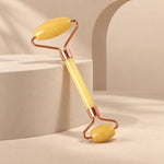 A yellow jade facial roller with rose-gold frame is angled on a light beige background with geometric blocks.