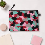 A flat, closed pouch with a pink, red, black, grey, and teal camouflage pattern, resting on a white desk alongside a