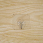 A transparent self-adhesive hook with a silver metal hook centered on a light wooden surface.