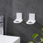Two white plastic bottle holders with clear adhesive pads lying on a light gray tiled floor beside a white bathtub.