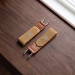 Two tan elastic belt hooks with brown faux leather accents and silver-colored metal hooks, resting on a dark wooden surface.