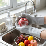 Hands in grey silicone scrubbing gloves wash a red apple over a kitchen sink with a bowl of fruit nearby.
