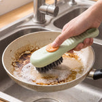 A hand uses a green and orange liquid-dispensing brush to clean a dirty frying pan in a stainless steel sink.