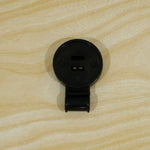 A single black plastic shade cloth clip, closed, lying flat on a light wood grain surface.