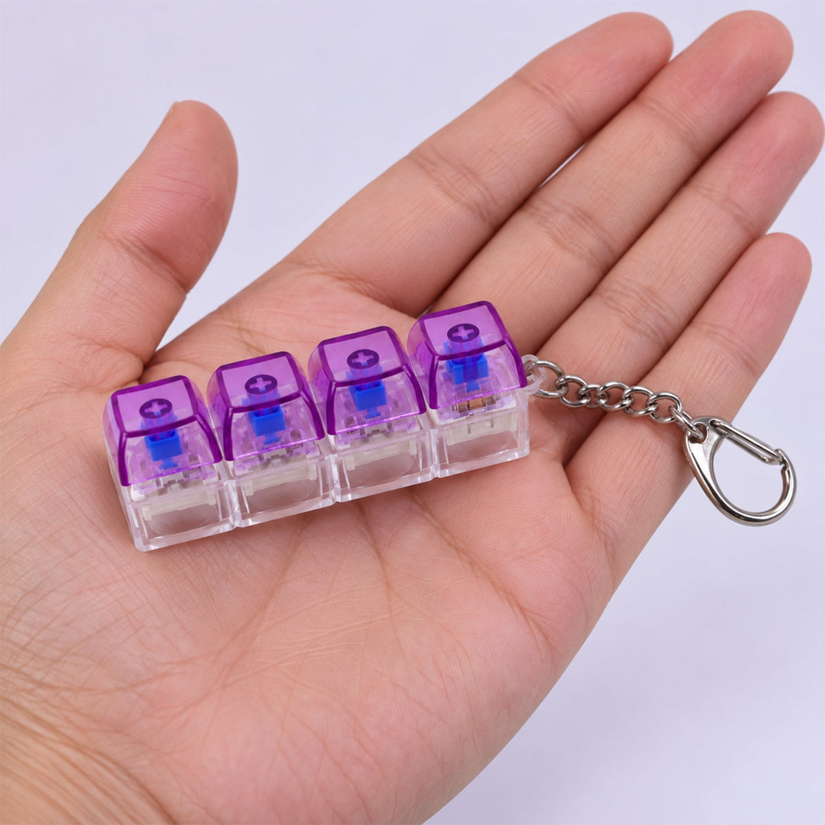 A four-key keyboard switch keychain with purple caps and a metal clasp held in the palm of a hand.